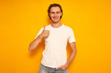 Gülümseyen kıvırcık saçlı, günlük kıyafetler giyen beyaz tişörtlü adam elini kaldırıyor, parmağını kaldırıyor, sarı arka planda soyutlanıyor. Metin için boşluk. İnsan konsepti, tavsiye, reklam