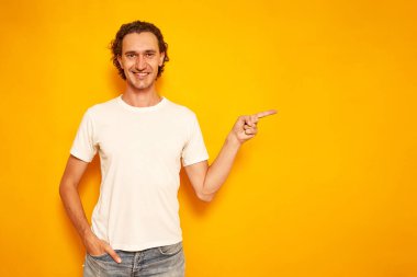 Gülümseyen kıvırcık saçlı, günlük kıyafetli bir adam parmağını reklam için boş bir yere doğrultuyor. Sarı arkaplanda izole edilmiş, mesaj için boşluk var. konsept insanları, promosyonlar, indirimler, tavsiyeler