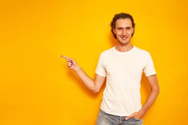 Mutlu, kıvırcık saçlı, sıradan giyinmiş bir adam reklam metni için parmağını boş yere doğrultuyor. Sarı arka planda izole edilmiş. İnsan terfileri, indirimler, reklam teklifleri, indeks kavramı