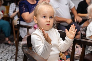 Sarışın, genç, beyaz bir kız sandalyeye oturup ellerini çırpıyor. O, uzun kollu beyaz bir gömlek giyiyor. Arka plan bir grup insanı gösteriyor..