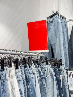Bir giyim mağazasında askıda asılı çeşitli kot pantolonlar. Arka planda parlak kırmızı bir tabela görülebiliyor, bu da satış veya promosyon anlamına geliyor.