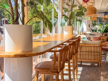 Ahşap masaları ve sandalyeleri olan modern bir restoran. Alan parlak ve havadar, dekorasyon için büyük pencereler ve bitkiler var..