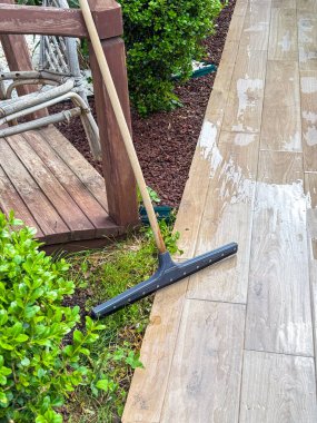 Yerde süpürgesi olan ıslak bir ahşap veranda. Kenarlarında yeşil çalılar ve arka planda kısmen görünen tahta bir sandalye var..