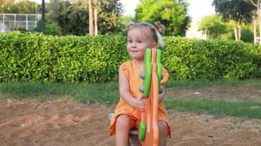 Sarışın, beyaz bir kız çocuk parkında tahterevalli gibi oturuyor. Yeşil çalılar ve çimlerle çevrili turuncu bir elbise giyiyor ve gülümsüyor..