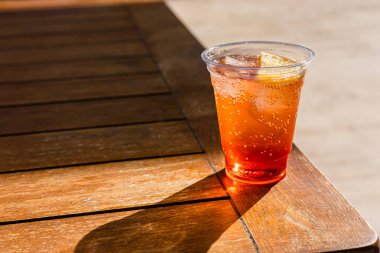 Buz küpleri ve bir dilim portakallı temiz plastik bardakta ferahlatıcı bir alkol kokteyli. İçki, yumuşak ışıklandırmalı ahşap bir masaya konur.. 