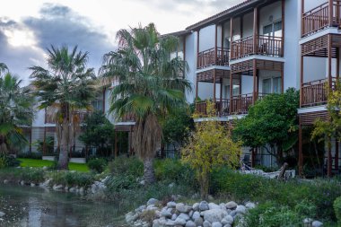 Bulutlu bir gökyüzünün altında sakin bir göletin yakınındaki palmiye ağaçları ve yeşilliklerle çevrili modern otel binası