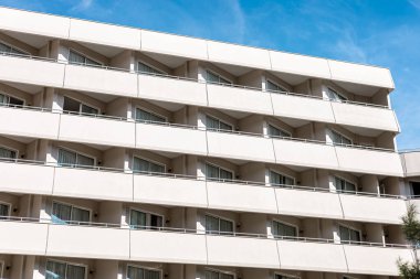 Çoklu balkonları ve geniş pencereleri olan modern bir apartman. Ön cephe açık renkli, açık mavi gökyüzüne karşı.