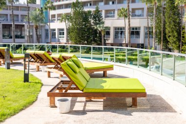 Yeşil yastıklı şezlonglar yüzme havuzunun yanındaki verandada düzenlenir. Palmiye ağaçları ve modern bir otel çevresi rahatlatıcı bir atmosfer yaratır.