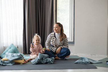 Uzun kahverengi saçlı beyaz bir kadın, küçük bir kızla birlikte paspasın üzerinde oturuyor. Kızın sarı saçları var ve oyuncaklarla oynuyor. Sıcak bir yuva ortamında battaniyelerle çevrili..