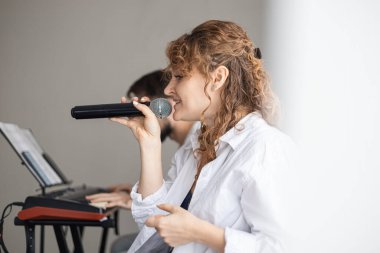 Kıvırcık saçlı beyaz bir kadın mikrofona şarkı söylüyor. Arka planda bir adam klavye çalıyor. Sahne bir ev konseri atmosferi yakalıyor..