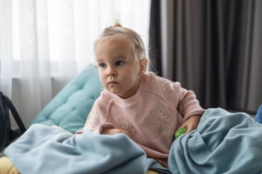 Açık kahverengi saçlı beyaz bir kız mavi bir battaniyeye sarılı bir yatakta oturuyor. Düşünceli bir şekilde pencereden dışarı bakıyor, rahat bir ev ortamı yaratıyor..