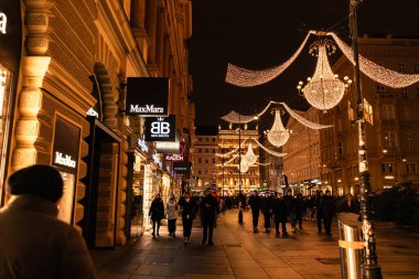 Viyana, Avusturya - 15 Aralık 2025: Avusturya 'nın Viyana kentindeki işlek cadde festival ışıklarıyla aydınlatıldı. Parıldayan avizeler dükkanların üzerinde asılı. Farklı geçmişlere sahip insanlar kışın yürürler..