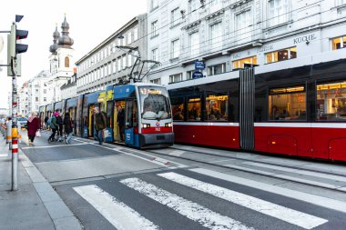 Viyana, Avusturya - 15 Aralık 2025: Viyana, Avusturya. Durakta mavi bir tramvay ve kırmızı bir tramvay park halindedir. Beyaz bir adam tramvayların yanından geçiyor..
