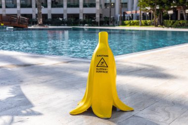 Sarı muz şeklinde uyarı levhası bir yüzme havuzunun yanında duruyor. Havuz modern binalar ve palmiye ağaçları ile çevrili, bu da tropikal bir ortamı gösteriyor.