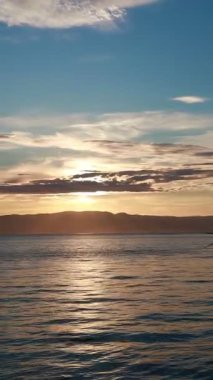 Sakin suların üzerinde sakin bir günbatımı, yumuşak dalgalar gökyüzünün altın ve mavi renklerini yansıtıyor. Dağlar uzaktan görülebilir..