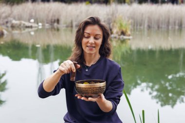 Kahverengi saçlı beyaz bir kadın dingin bir gölün kenarında elinde renkli malzemelerle dolu ahşap bir kâse tutuyor. Suyu çevreleyen yeşil sazlıklar.