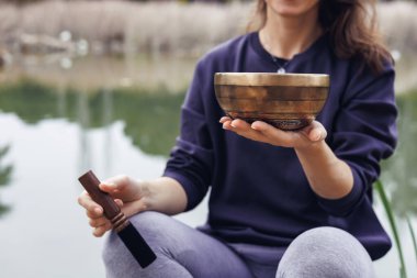 Uzun kahverengi saçlı beyaz bir kadın dingin bir gölün yanında ahşap bir kase tutuyor. Arka planda ağaçlar ve yeşillikler görülebilir..