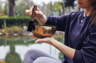 Kahverengi saçlı genç bir kadın, sakin bir su kütlesinin yanında açık havada şarkı söylüyor. Koyu renk bir kazak ve gri tayt giyer. Rahatlamayı ve farkındalığı teşvik eder..