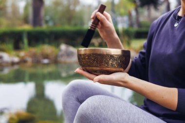 Kahverengi saçlı genç bir kadın, sakin bir su kütlesinin yanında açık havada şarkı söylüyor. Koyu renk bir kazak ve gri tayt giyer. Rahatlamayı ve farkındalığı teşvik eder..