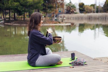 Uzun kahverengi saçlı beyaz bir kadın gölün kenarındaki yeşil yoga minderinde bacak bacak üstüne atmış oturuyor. Elinde bir kaseyle meditasyon yapıyormuş gibi görünüyor..