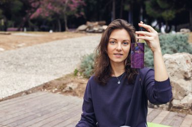 Kahverengi saçlı, gölün kenarında oturan, elinde akıllı telefon tutan beyaz bir kadın. Koyu renk bir kazak ve gri pantolon giyiyor, dışarıda huzurlu bir anın tadını çıkarıyor..