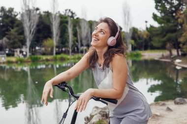 Kahverengi saçlı ve pembe kulaklıklı genç beyaz kadın parkta sakin bir gölün kenarında elektrikli scooter 'a yaslanırken gülümsüyor..