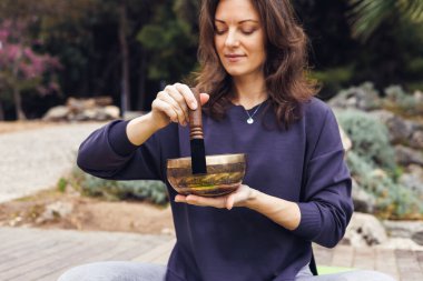Kahverengi saçlı, beyaz bir kadın açık havada şarkı söylüyor. Koyu renk bir kazak giyer ve bahçedeki bir paspasın üzerine oturur..