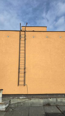 Açık turuncu bir binanın duvarında dikey metal bir merdiven. Aşağıdan yukarıya bakıldığında, temiz geometri, minimalizm ve endüstriyel estetik içeriyor..