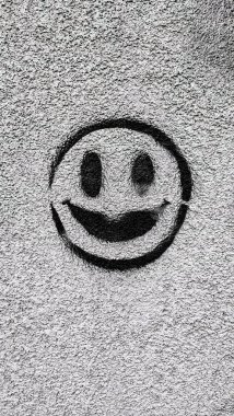 Açık gri, sert beton bir duvarda gülümseyen bir yüzün minimalist siyah graffitisi. Sokak tarzı, desenli yüzey, kumlu doku. Beton duvardaki gülen surat duvar yazısı. Minimalist sokak sanatının arka planı.