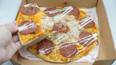 Bir el pizza kutusundan sosis ve mozzarella peynirli bir dilim pizza alıyor.