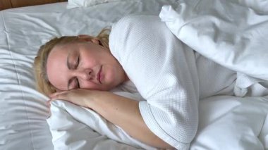 Yatakta beyaz çarşaflarla uyuyan bir kadın. Görüntü, rahat bir yatak odası ortamında huzurlu ve huzurlu bir anı yakalıyor. Rahatlama, uyku ve rahatlama kavramları için idealdir. Yüksek kalite 4k