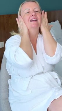 Bornozlu olgun sarışın kadın yatağın yanındaki sandalyeye oturur, nazikçe yüzüne masaj yapar ve krem sürerek, rahat yatak odasında kendi kendine bakım ayini yapar..