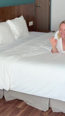 Neşeli yetişkin bir kadın beyaz bir kanepeye uzanmış, gülümsüyor ve telefonda sohbet ediyor. Balkon penceresinden gelen güneş ışığı evin içini doldurur..