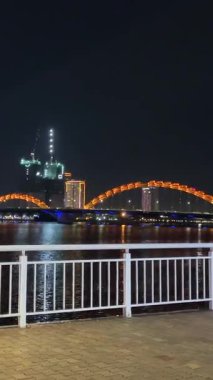 Han Nehri 'nin üzerinde, Da Nang' daki Ejderha Köprüsü 'nün parlak turuncu manzarası. Şehir Vietnam 'ın karanlık gökyüzünün altında parlarken yansımalar sakin sularda dans ediyor.. 