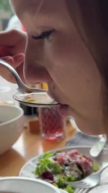 Beyaz tabaktan çorba kepçeleyen ve pencere kenarındaki restoran masasına kaşıkla yemek getiren dışarıda bulanık insanlar ve arabalar ve öğle yemeği sırasında yakınlarda aile yemeği yiyen bir kadın.. 