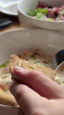 Kadın ekmeğe karides koyup parmaklarıyla uyum sağlamak için çatal kullanırken aile yemeğinde kremalı sosu beyaz tabaklar ve taze yiyeceklerle birlikte ahşap masaya sürüyor..