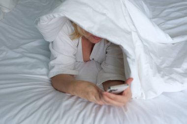 Cep telefonunu kullanırken başının üzerinde battaniyeyle yatakta uzanmış gülümseyen bir kadın. Yüksek kalite 