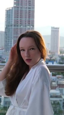 Beyaz bornozlu neşeli genç bir kadın güneşli bir balkonda duruyor. Parlak, modern bir otelde sabah özgürlüğünün ve rahatlığının tadını çıkararak uzun saçlarını düzeltiyor..