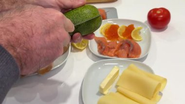 Sarı tişörtlü kızıl saçlı kadın dışarıda cam masada oturuyor, gri kesme tahtasına somon ve avokado dilimliyor, ekmek ve krem peynirle sağlıklı bir yemek hazırlıyor. Yüksek kalite 4k görüntü