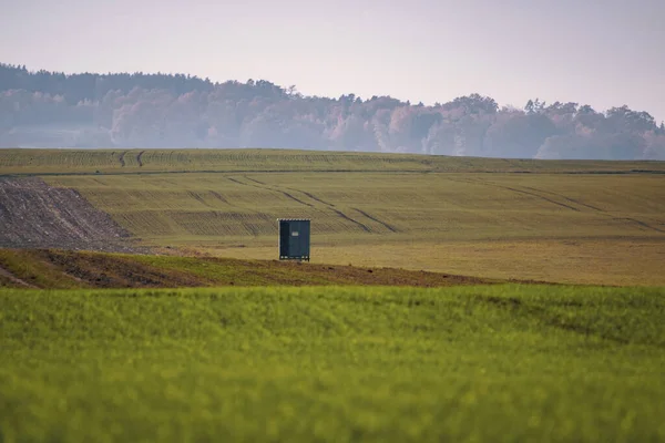 Puslu bir sonbahar gökyüzünün altında yapayalnız bir av durağı, ekilmiş tarım arazileri ve uzak orman tepeleriyle çevrili, yalnızlığı ve huzurlu kırsal atmosferi sembolize eden,.