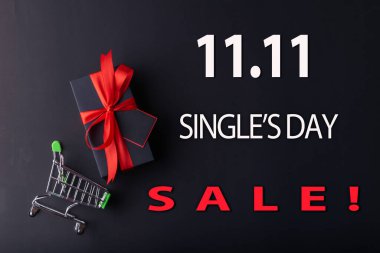 11.11 Bekar Günü indirimi. Siyah arka planda kırmızı kurdeleli kırmızı hediye ve siyah arka planda küçük el arabası ile beyaz 11.11 single 'ın günlük satışı metni