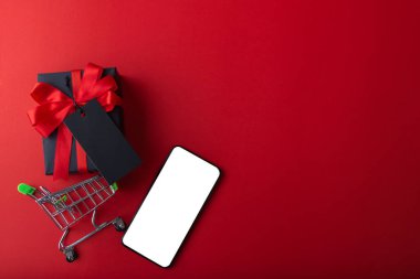 Kırmızı arka planda siyah etiketli siyah bir hediye ve kırmızı kurdele, küçük el arabasında siyah kredi kartı ve alışveriş için akıllı telefon. 11.11 bekarlar günü indirimi, Pazartesi indirimi veya Kara Cuma indirimi konsepti.