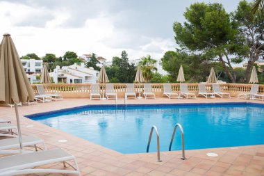 Mavi yüzme havuzu. Etrafta bir sürü güneşlik ve şemsiye var. Mallorca adasında yeşil ağaçlar ve villalarla çevrili dinlenme bölgesi.