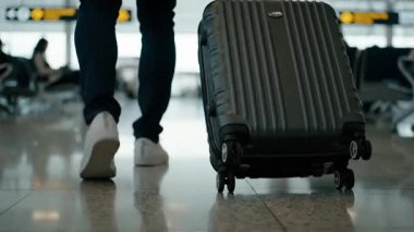 Bir gezgin havaalanı terminalinden elinde sert bir bavulla geçiyor. Düşük açılı çekim, arka planda bulanık yolcular ve kapı işaretleriyle cilalı zeminde yuvarlanan bagaj tekerleklerine odaklanıyor..