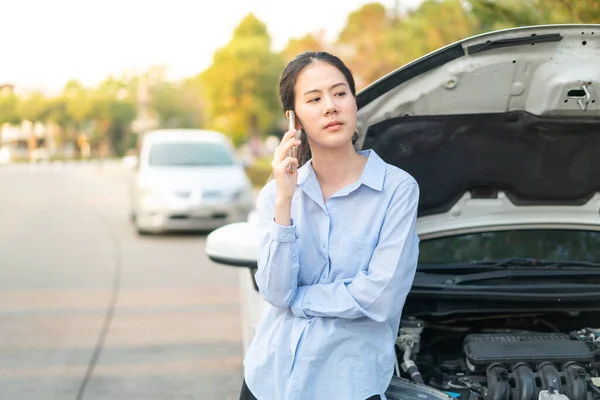 Young Asian woman standing near broken down car with popped up hood having trouble with her vehicle