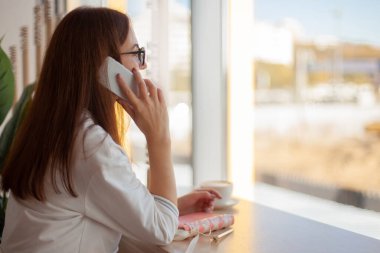 Genç bayan telefonda konuşuyor ve sağlıklı sıcak kahve içerken notlar yazıyor. Güzellik ve refah