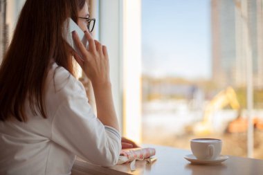 Genç bayan telefonda konuşuyor ve sağlıklı sıcak kahve içerken notlar yazıyor. Güzellik ve refah