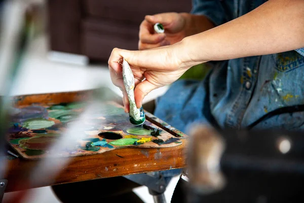 Woman artist hand squeezing a tube with yellow orange oil paint on wooden palette for painting in art studio. Concept of creating new colours by mixing oil paint. Unfocused paintbrushes.