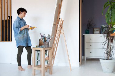 Boyalı fırça tutan ve yağlı boyayla resim yapan genç bir kadın ressam. Çerçevelenmiş pamuk tuval üzerinde evdeki sehpada. Hobi ve eğlence konsepti. Ressam sanat yaratıyor. İç mekan. Boşluğu kopyala.
