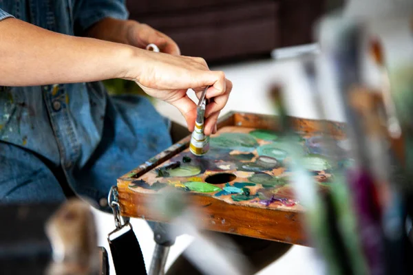Woman artist hand squeezing a tube with yellow orange oil paint on wooden palette for painting in art studio. Concept of creating new colours by mixing oil paint. Unfocused paintbrushes.